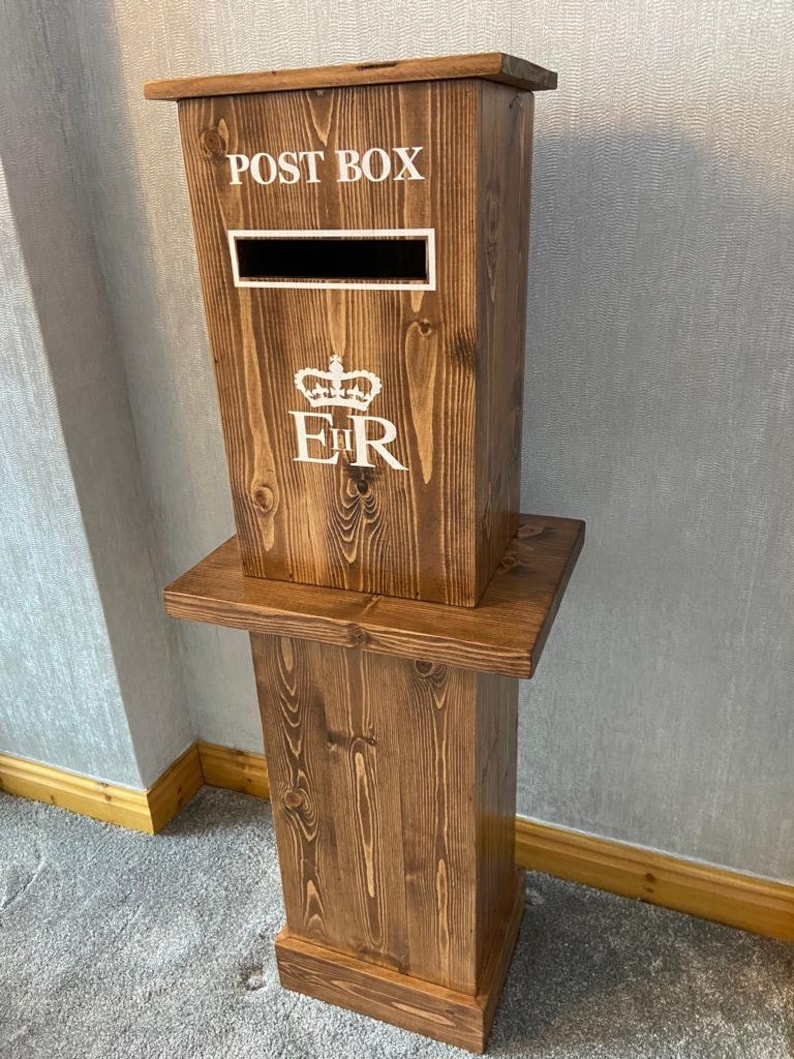 May include: A wooden post box with a royal crest and the words "POST BOX" on the front. The post box is on a wooden stand.