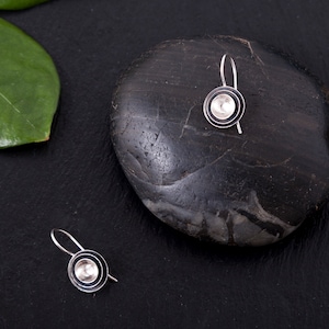 May include: A pair of silver earrings with a circular design. The earrings are on a black surface with a gray rock.