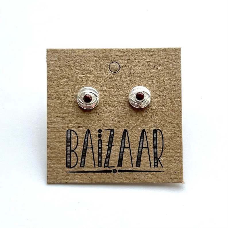 May include: A pair of silver earrings with a red eye design. The earrings are on a brown card with the word "BAIZAAR" printed on it.