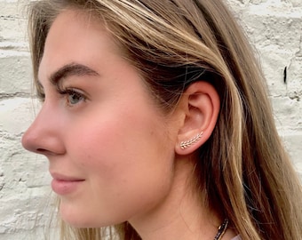 May include: A woman wearing a gold leaf ear cuff earring. The earring is a delicate, thin piece of jewelry that is shaped like a leaf. The earring is worn on the upper part of the ear, just above the lobe.