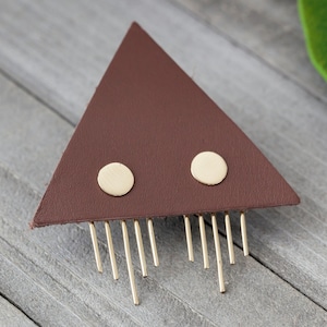 May include: A brown leather triangle with two gold circles and a fringe of gold metal strands.