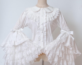 May include: White blouse with a Peter Pan collar, ruffled trim, and long sleeves with flared, lace-trimmed cuffs. The blouse features a button-down front and is displayed on a mannequin. The sleeves have bows at the elbow.