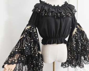 May include: Black off-the-shoulder blouse with long, flared sleeves. The blouse has a ruffled neckline and cuffs, with intricate lace and gold detailing. The sleeves are made of delicate, patterned lace. The blouse is displayed on a mannequin.