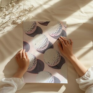 May include: A print featuring golf balls with a pink and purple background. The golf balls are white with dimpled surfaces, and the print is held by hands. The print is on a cream-colored surface with shadows.