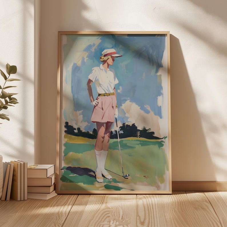 May include: A framed vintage-style golf art print. The artwork depicts a person in a white shirt, pink skirt, and golf attire, holding a golf club on a green course. The background features a blue sky with clouds and a green landscape.