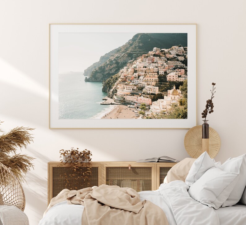 May include: A framed photograph of a coastal town built into a hillside. The town is a mix of white and pastel-colored buildings with a few trees and a small beach in the foreground. The sea is a light blue color and the sky is a pale blue.