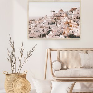 May include: A framed print of a whitewashed village with a windmill on a hill overlooking the ocean. The buildings are painted in shades of white, pink, and yellow. The print has a rustic, vintage feel.
