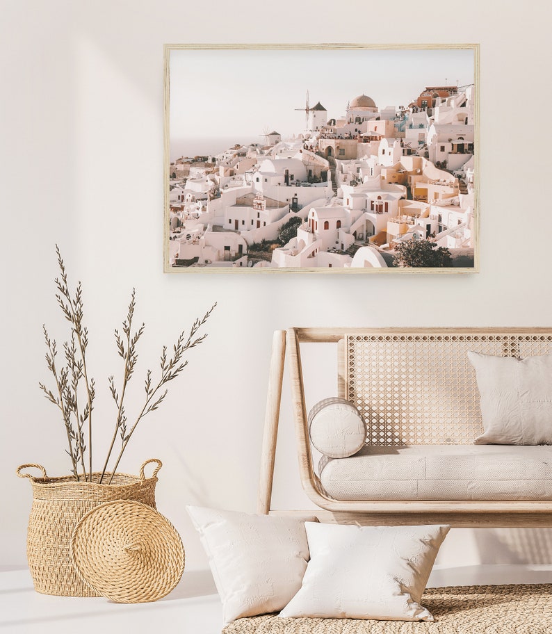 May include: A framed print of a whitewashed village with a windmill on a hill overlooking the ocean. The buildings are painted in shades of white, pink, and yellow. The print has a rustic, vintage feel.