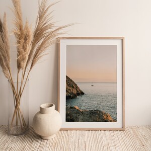 May include: A framed photograph of a seascape with a sailboat in the distance. The photo is in a light wood frame and is hanging on a white wall. There are dried pampas grasses in a clear vase on a woven rug in front of the photo.