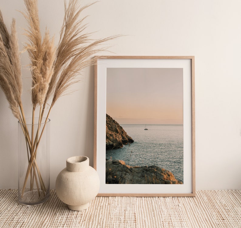 May include: A framed photograph of a seascape with a sailboat in the distance. The photo is in a light wood frame and is hanging on a white wall. There are dried pampas grasses in a clear vase on a woven rug in front of the photo.
