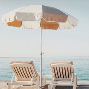 Puede incluir: Dos tumbonas blancas con cojines beige dan a la playa bajo una sombrilla de playa a rayas blancas y beige.