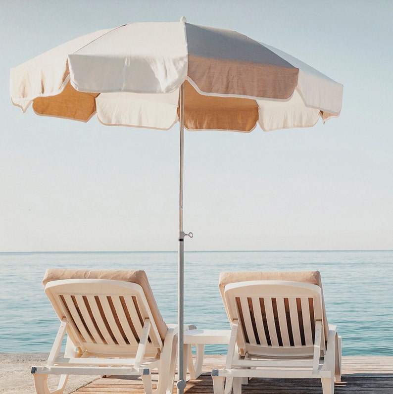 Puede incluir: Dos tumbonas blancas con cojines beige dan a la playa bajo una sombrilla de playa a rayas blancas y beige.