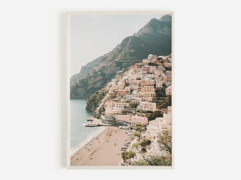 May include: A photograph of a coastal town built into a hillside, with a sandy beach and clear blue water in the foreground. The town is a mix of white and pastel-colored buildings, with lush green vegetation growing on the hillside.