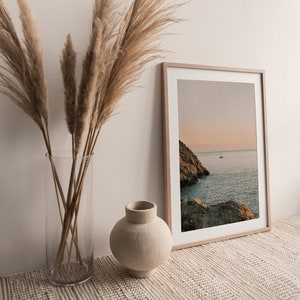 May include: A framed photograph of a rocky coastline with a sailboat in the distance. The photo is in a light wood frame and is sitting on a beige rug. A vase with dried pampas grass is in the foreground.