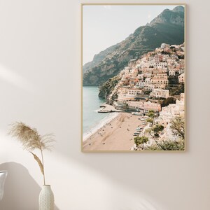 May include: A photograph of a colorful town built on a cliffside overlooking a sandy beach and the ocean. The town is a mix of pastel colors, with white buildings and terracotta roofs. The beach is empty, with a few people walking along the shore.