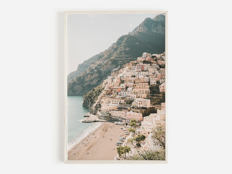 May include: A photograph of a coastal town built into a hillside, with a sandy beach and clear blue water in the foreground. The town is a mix of white and pastel-colored buildings, with lush green vegetation growing on the hillside.