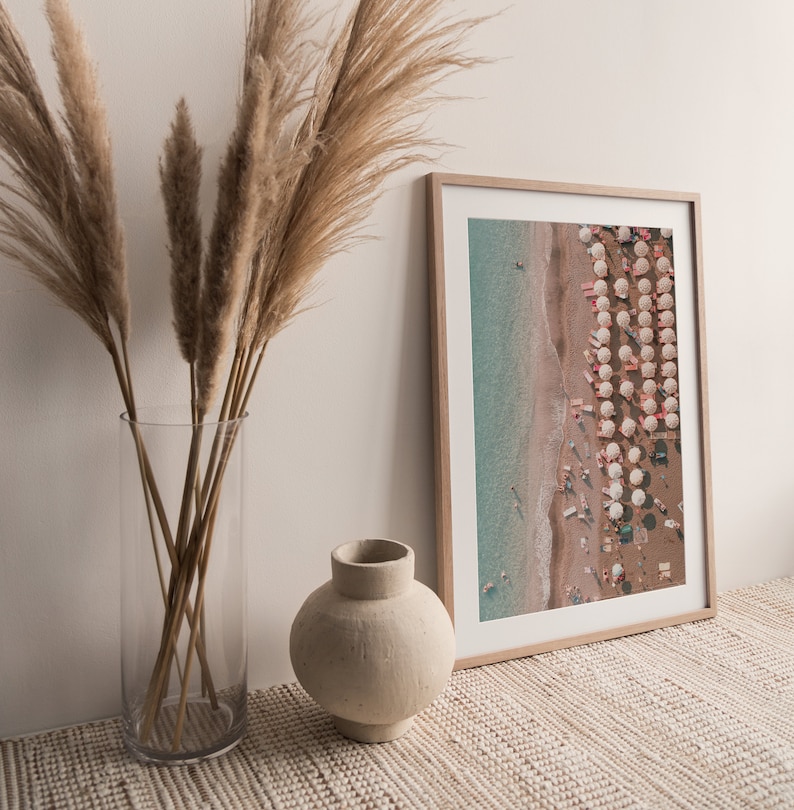 Peut inclure: Une photographie encadr&eacute;e d'une sc&egrave;ne de plage avec des parasols blancs et des gens dans l'eau. La photo est accroch&eacute;e &agrave; un mur blanc avec un vase de pampas s&eacute;ch&eacute;es devant.