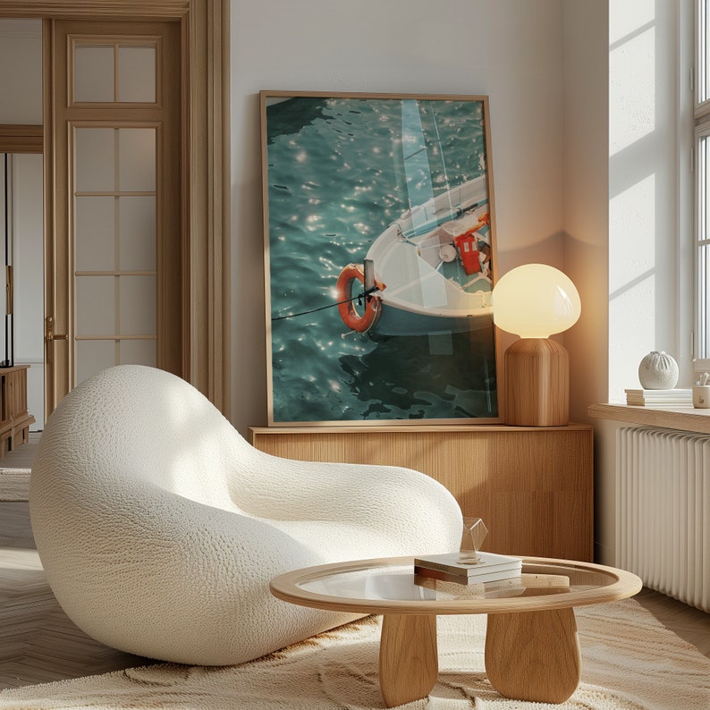 Op de afbeelding: Een witte, pluche fauteuil met een houten salontafel ervoor. Een ingelijst afdruk van een boot op het water hangt aan de muur achter de fauteuil. Een lamp met een witte kap staat op een houten plank.