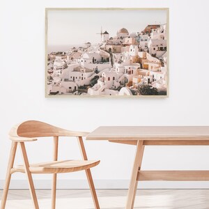 May include: A framed photograph of a whitewashed village with a windmill on a hilltop. The village is built on a cliff overlooking the sea. The photo is in a light wood frame.