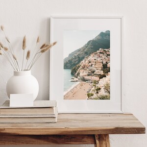 May include: A white framed photograph of a colorful town built on a hillside overlooking a beach. The town is surrounded by green hills and a blue ocean.