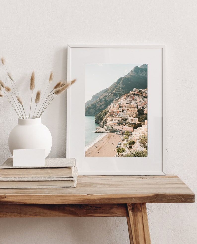 May include: A white framed photograph of a colorful town built on a hillside overlooking a beach. The town is surrounded by green hills and a blue ocean.