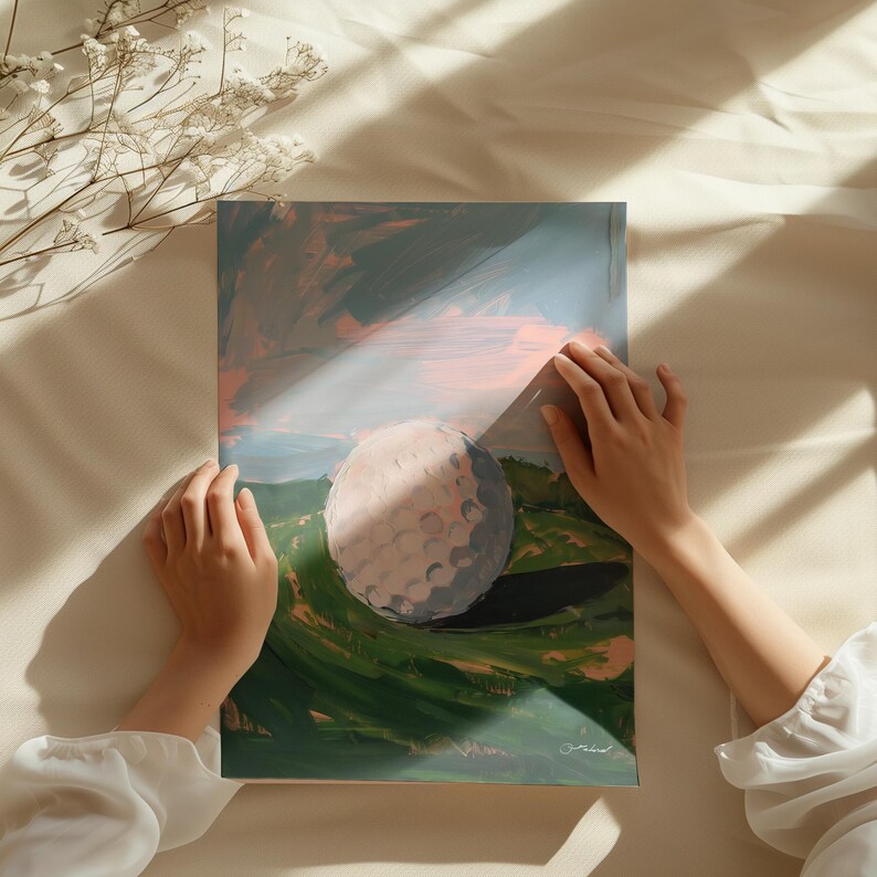 May include: An abstract painting of a golf ball on a green landscape under a blue and pink sky. The artwork is held by two hands, with a soft, neutral background. The painting has a textured, artistic style.