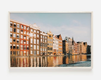 Könnte beinhalten: Eine Reihe von bunten Backsteingebäuden mit Bogenfenstern und roten Ziegeldächern, die sich in einem Kanal spiegeln. Die Gebäude stehen am Rande des Wassers, und die Sonne scheint hell am Himmel.