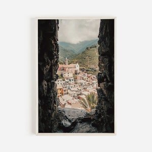May include: A view of a small village nestled in a valley, seen through a stone window. The village is made up of colorful buildings with terracotta roofs, and the valley is covered in lush green vegetation.