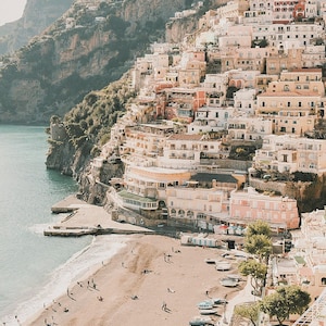 May include: A view of a colorful town built on a cliffside overlooking a sandy beach with turquoise water. The town is made up of many buildings with terracotta roofs and white walls. There are people on the beach and boats in the water.