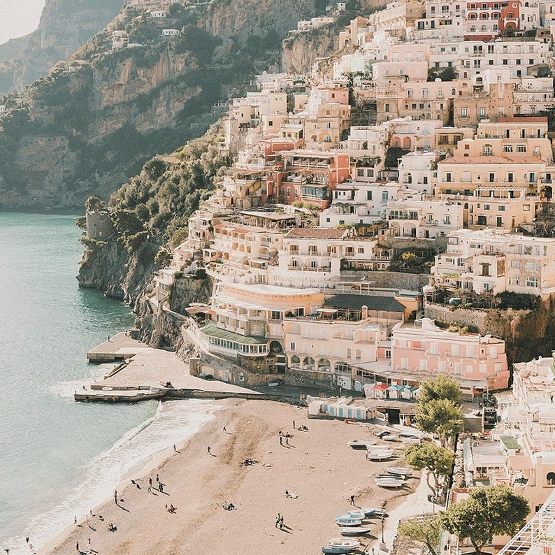 May include: A view of a colorful town built on a cliffside overlooking a sandy beach with turquoise water. The town is made up of many buildings with terracotta roofs and white walls. There are people on the beach and boats in the water.