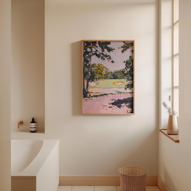 May include: A framed landscape painting depicting a golf course scene with trees, a green fairway, and a pinkish path. The artwork is in a light wooden frame and hangs on a neutral-colored wall. A woven basket sits on the floor below.