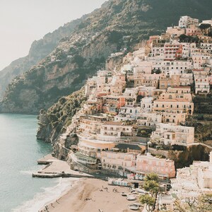 May include: A view of a town built on a cliffside overlooking a sandy beach and the ocean. The town is made up of colorful buildings with terracotta roofs. The sky is a light blue and the water is a deep blue.