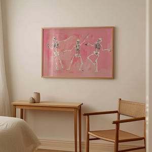 May include: Three skeletons dancing on a pink background in a framed print. The skeletons are in various poses, with their arms and legs outstretched. The background is a soft pink with a watercolor effect.
