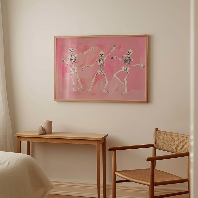 May include: Three skeletons dancing on a pink background in a framed print. The skeletons are in various poses, with their arms and legs outstretched. The background is a soft pink with a watercolor effect.