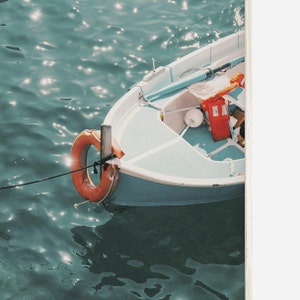 Op de afbeelding: Een klein wit bootje met een blauw interieur ligt aangemeerd in het water. Een feloranje reddingsboei is aan de zijkant van het bootje bevestigd. Het water heeft een levendige blauwgroene kleur.