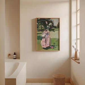 May include: A framed painting of a golf course scene. A pink golf bag with clubs is on green grass, with trees in the background. The artwork is in a light wooden frame, complementing the room's decor.