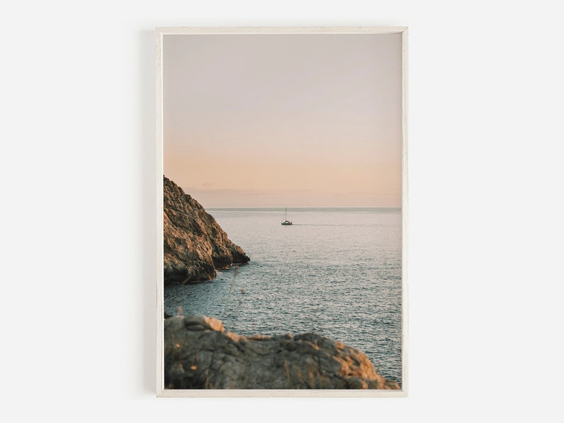 May include: A photograph of a rocky cliff overlooking a calm ocean with a single sailboat in the distance. The sky is a soft pink and purple, creating a serene and peaceful atmosphere.