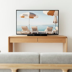 Puede incluir: Un mueble de TV de madera con dos cajones, un televisor de pantalla plana que muestra una escena de playa con tumbonas y sombrillas, y un sof&aacute; gris en primer plano.