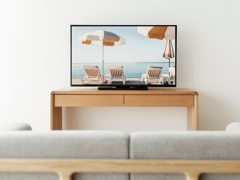 Puede incluir: Un mueble de TV de madera con dos cajones, un televisor de pantalla plana que muestra una escena de playa con tumbonas y sombrillas, y un sof&aacute; gris en primer plano.