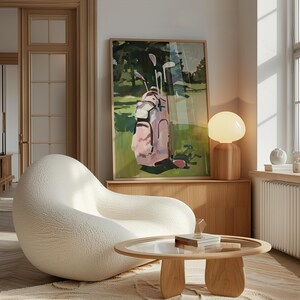 May include: A framed art print showing a golf bag with clubs on a green golf course. The golf bag is pink and white. The artwork is displayed above a wooden cabinet, next to a white, rounded chair and a round wooden coffee table.