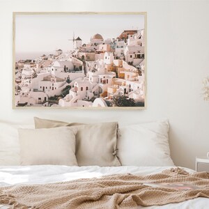 May include: A framed photograph of a whitewashed village with a windmill on a hilltop. The buildings are clustered together and have a rustic, charming feel. The sky is a soft blue and the sun is shining.