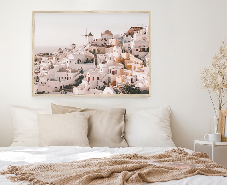 May include: A framed photograph of a whitewashed village with a windmill on a hilltop. The buildings are clustered together and have a rustic, charming feel. The sky is a soft blue and the sun is shining.