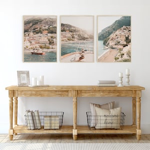 May include: A wooden console table with two shelves, featuring three framed photographs of a coastal town with buildings and a boat in the water. The table has two black wire baskets on the bottom shelf, one with books and the other with a pillow and the text "welcome home".