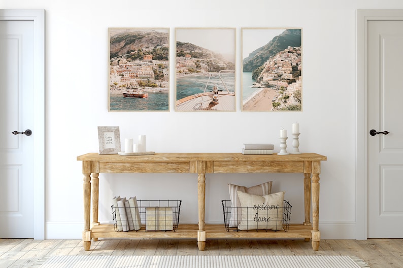 May include: A wooden console table with two shelves, featuring three framed photographs of a coastal town with buildings and a boat in the water. The table has two black wire baskets on the bottom shelf, one with books and the other with a pillow and the text "welcome home".