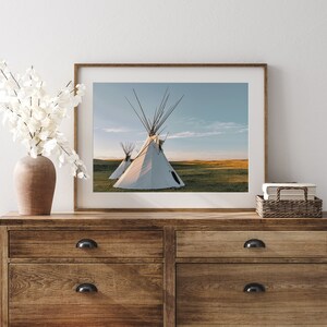 May include: A framed photograph of two white teepees on a grassy field with a blue sky in the background. The teepees are in the foreground of the image, and the sky is in the background. The image is framed in a light wood frame.