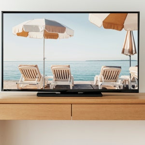 Puede incluir: Un televisor que muestra una escena de playa con tres tumbonas bajo sombrillas. El televisor est&aacute; sobre una mesa de madera.
