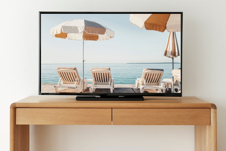 Puede incluir: Un televisor que muestra una escena de playa con tres tumbonas bajo sombrillas. El televisor est&aacute; sobre una mesa de madera.