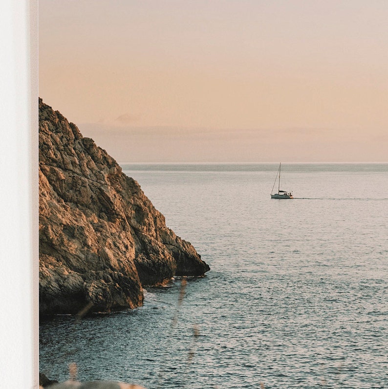 May include: A sailboat on a calm, blue ocean with a rocky cliff in the foreground. The sky is a soft pink and orange.