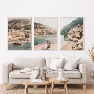 May include: Three framed photographs of a coastal town with colorful buildings on a hillside. The middle photo shows a boat in the water with the town in the background. The photos are hung on a white wall above a light beige sofa.