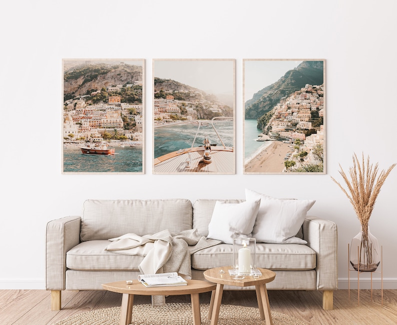 May include: Three framed photographs of a coastal town with colorful buildings on a hillside. The middle photo shows a boat in the water with the town in the background. The photos are hung on a white wall above a light beige sofa.